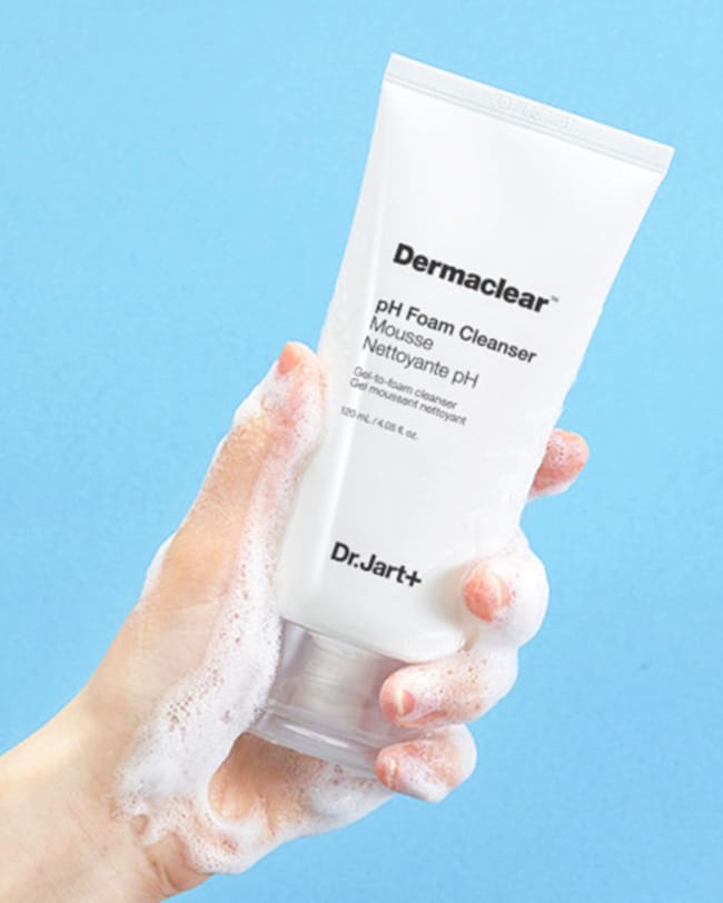 A hand holds up a bottle of Dermaclear Face Wash Cleanser as a frothy, cleanser, face wash flows over the tube and hand. 
