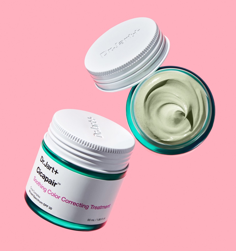 Two jars of color correcting cream are floating on a light pink background. One bottle is open to show the green cream texture. 