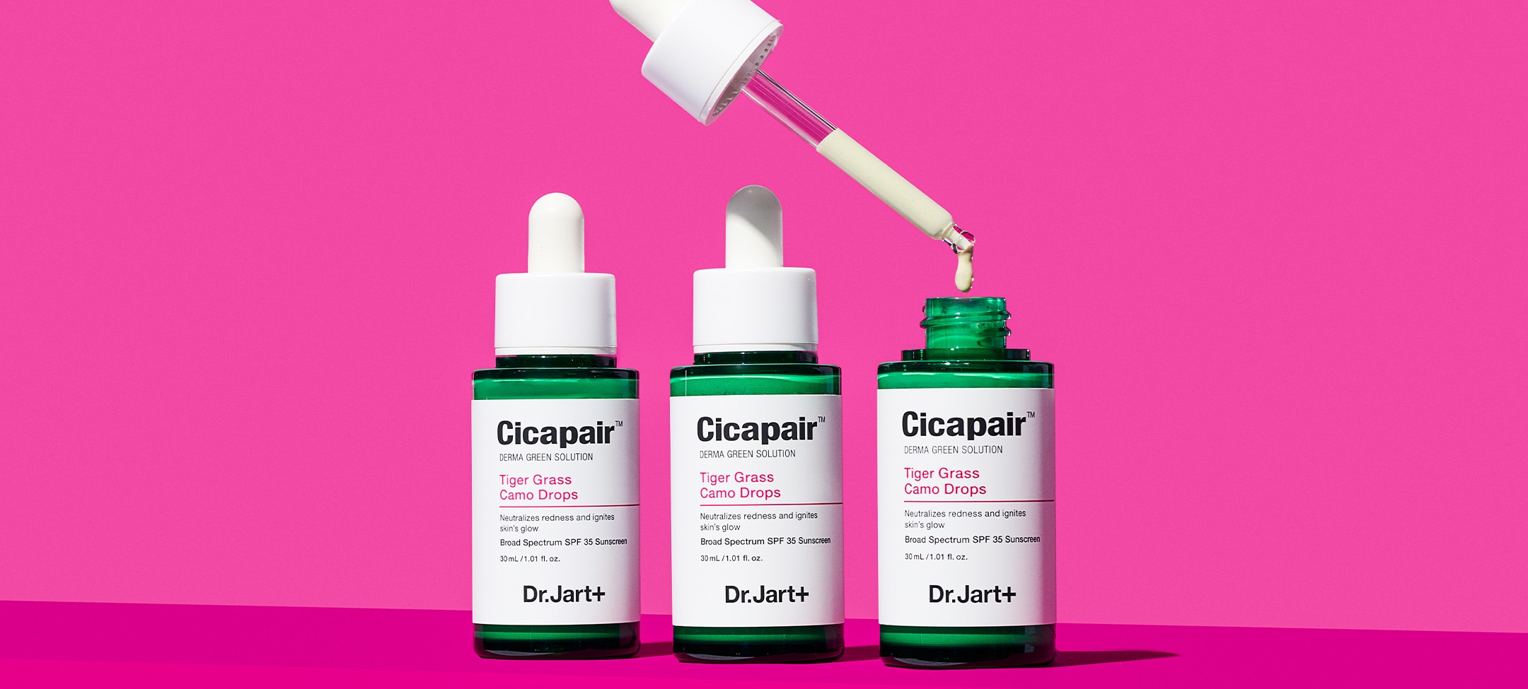 Three bottes of Camo Drops are displayed on a pink background with one applicator dropping the texture into an open bottle
