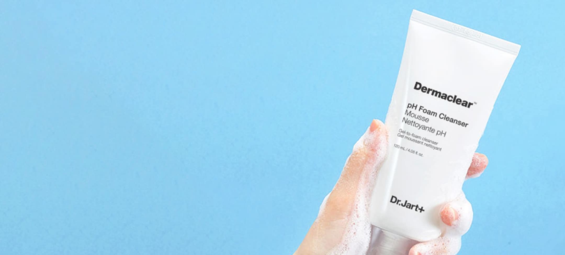 A hand holds up a bottle of Dermaclear Face Wash Cleanser as a frothy, cleanser, face wash flows over the tube and hand. 