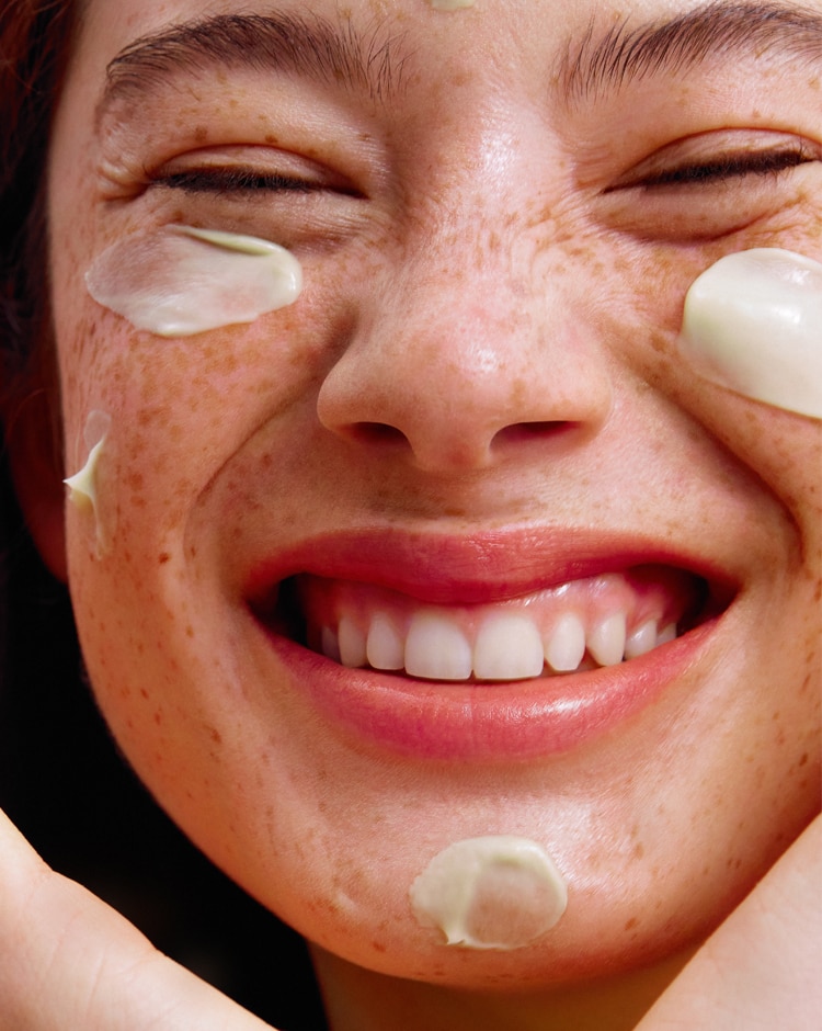 Woman smiles with dollops of Cicapair moisturizer cream applied to her cheeks and chin.